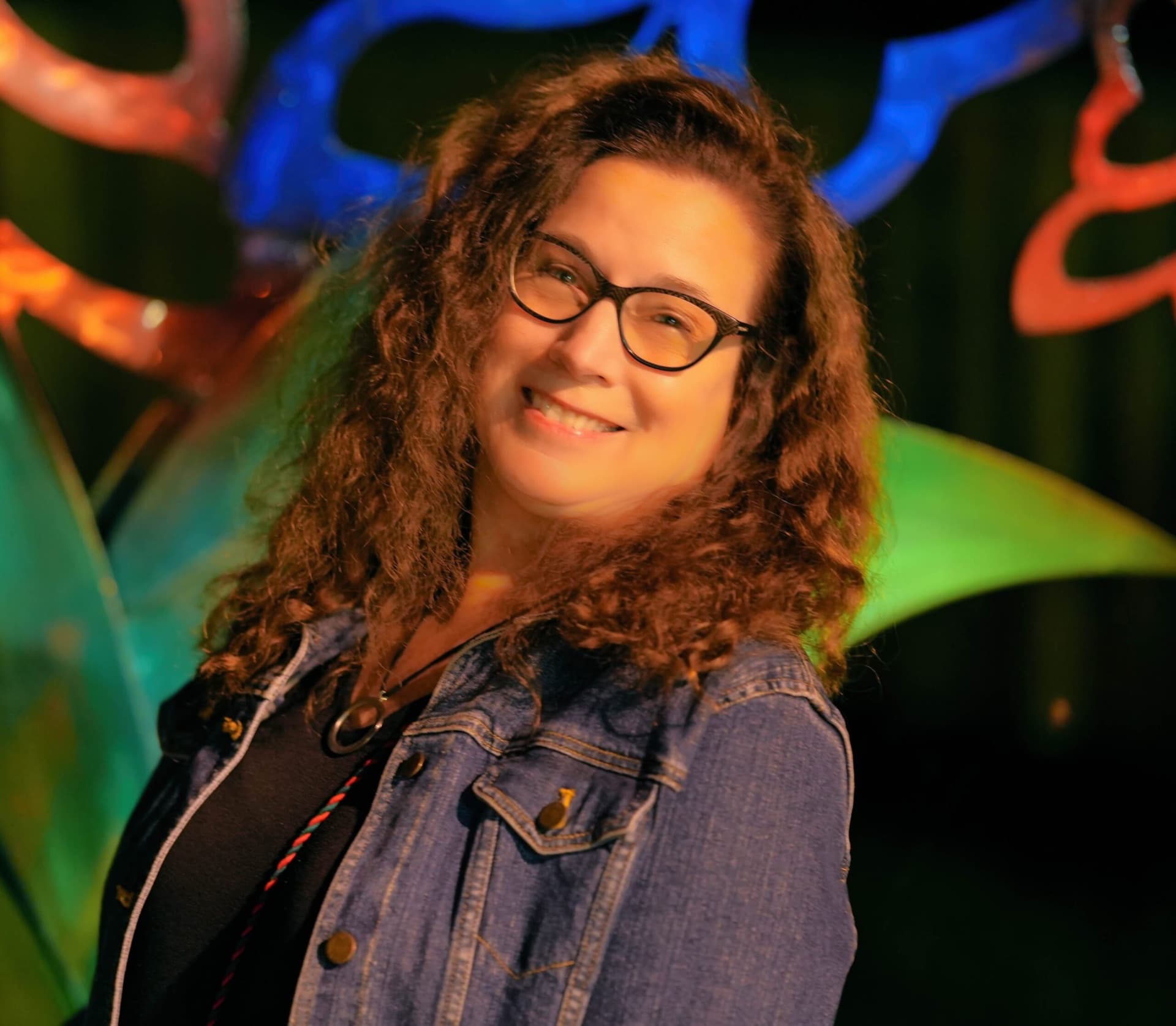 Night portrait of a smiling woman in a denim jacket against colorful neon sculpture light