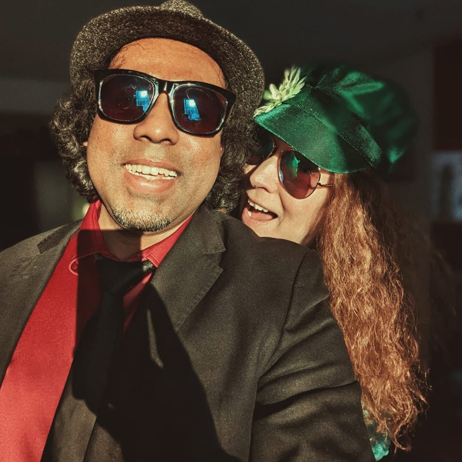 Couple in party outfits — fedora, shades and an emerald hat — laughing at a celebration
