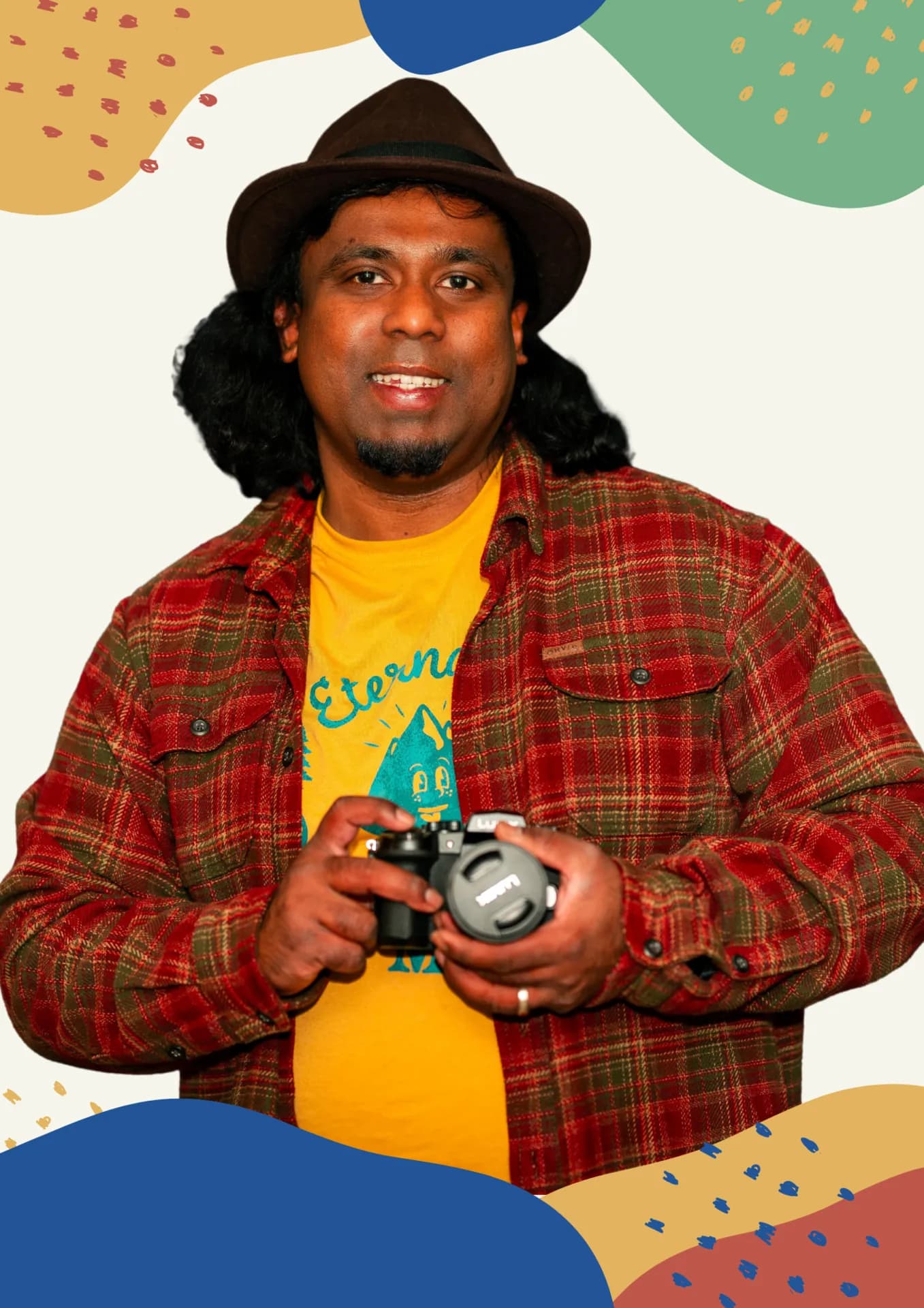 Jose Sharvin in a fedora and flannel shirt, smiling and holding a mirrorless camera